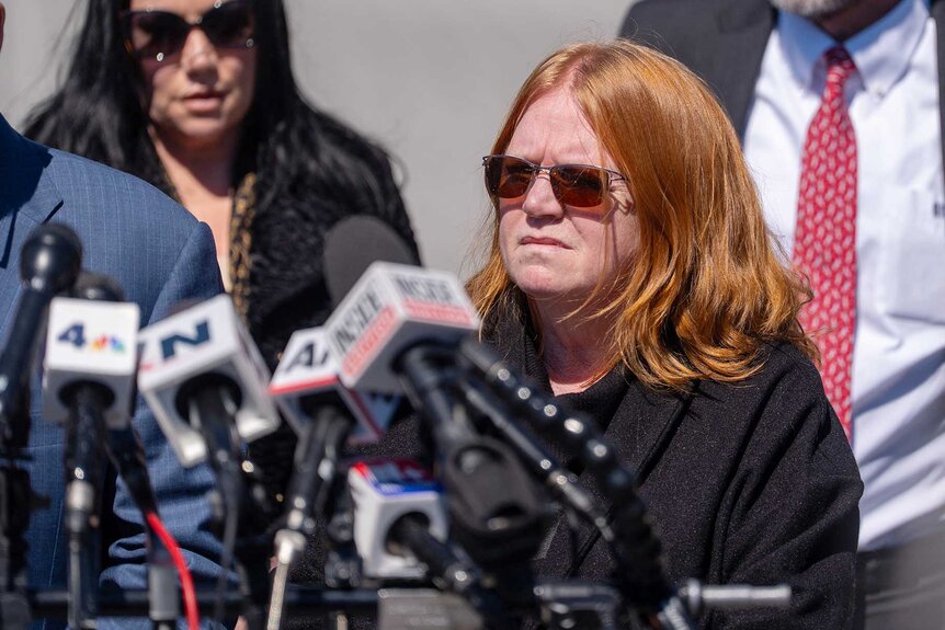 Asa Ellerup listening to Attorney Robert Macedonio speak in front of a group of microphones.
